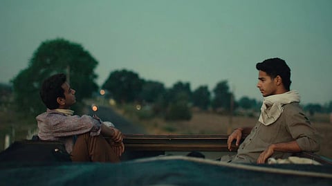 Actor Ishan Khatter and Vishal Jethwa sit facing each other on top of a vehicle at dusk, surrounded by an open rural landscape. Their expressions are thoughtful and contemplative.
