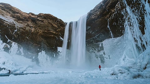 A person in a red jacket stands on ice near a majestic frozen waterfall, surrounded by snow-covered cliffs, evoking a sense of awe and solitude.