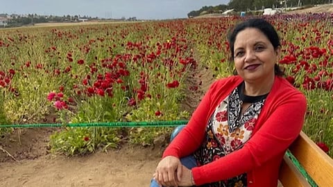 A woman in a red cardigan sits on a bench, smiling. Behind her is a vast field of vibrant red flowers under a cloudy sky, creating a serene atmosphere.