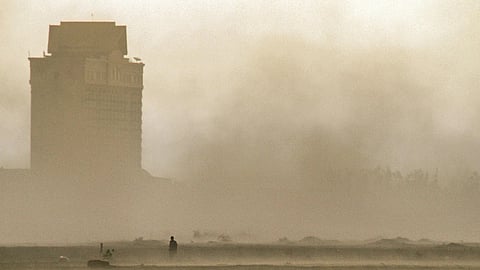 A lone figure stands in a vast, dusty field with sparse vegetation, shrouded in haze. A distant silhouette of a tall building looms, creating an eerie, desolate mood.