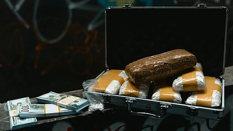 Open suitcase filled with plastic-wrapped packages, possibly drugs, beside stacks of $100 bills on a graffiti-covered surface.