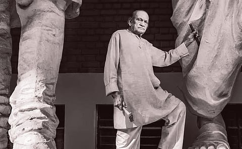 Ram Sutraj is shown in a beige kurta and white pants stands confidently between two large statues, holding sculpting tools, conveying creativity and pride.