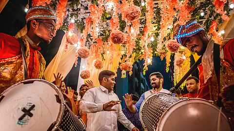 People celebrate joyfully under vibrant floral decorations and warm lights, with musicians in colorful attire playing large drums. 