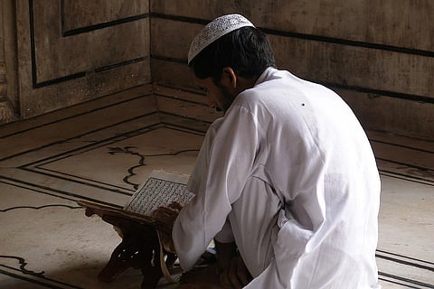 The image shows a Muslim person reading the Quran in Jama Masjid mosque. He is dressed in white kurta pyjama and white Muslim cap.