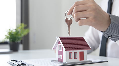 A hand in a formal shirt holds house keys above a model house on a contract, symbolizing real estate or home buying. A plant is blurred in the background.