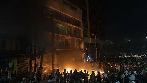A large crowd gathers at night around a fire outside a building, casting an orange glow. The scene conveys tension and unrest.