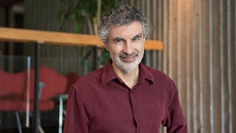 AI pioneer Yoshua Bengio with curly gray hair and a beard, wearing a maroon shirt, stands indoors. The background includes a red chair and vertical wooden panels.