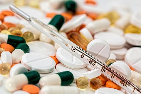 A pile of assorted pills, capsules, and gel tablets in various colors with a glass thermometer placed diagonally across them, conveying a medical theme.