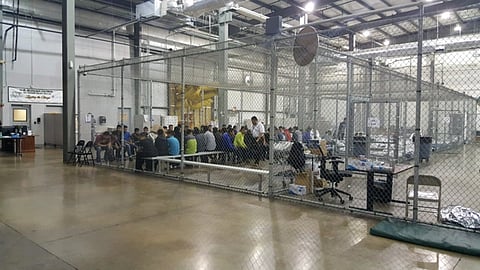 A detention center in Texas, USA. A large auditorium has a chainlink enclosure with many people sitting inside.