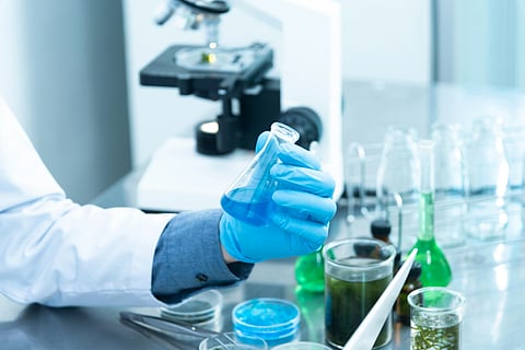 A scientist wearing blue gloves holds a flask with blue liquid in a lab. A microscope and several glassware items with colorful substances are on the table.