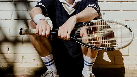 Man with a retro style sits on a tennis court, leaning against a white brick wall. He holds a racket, with two tennis balls at his feet.