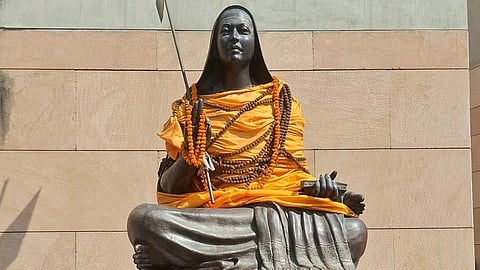 A bronze statue of a seated figure draped in vibrant orange cloth and adorned with garlands. The person holds a stick, exuding serenity and wisdom.