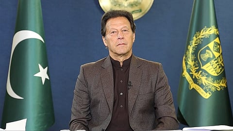 In the image Imran Khan is shown with a serious face looking at the camera behind him are two flag one of Pakistan and one prime minister flag