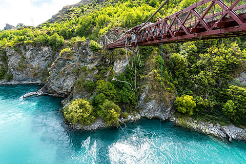 A person is bungee jumping from a rusty red bridge over a vibrant turquoise river, surrounded by lush green cliffs and trees.