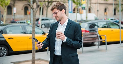 A man in a suit smiles while looking at his phone, holding a coffee cup. He stands on a city street, with taxis and a bench in the background.
