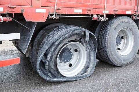 Close-up of a red truck with a severely damaged flat tire, its rubber shredded and hanging from the rim, on a wet road. The scene suggests an accident or mechanical failure.