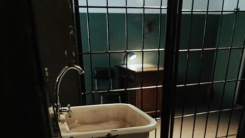Dimly lit prison cell with a sink and two dumbbells on the floor. Metal bars separate the space, casting shadows that convey a somber mood.