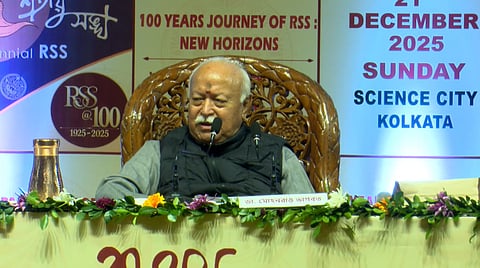 The image depicts RSS Chief Mohan Bhagwat addressing an event in Kolkata. Behind him, the posters depict "100 Years Journey of the RSS: New Horizons", and other things such as "21 December, 2025, Sunday, Science City, Kolkata".