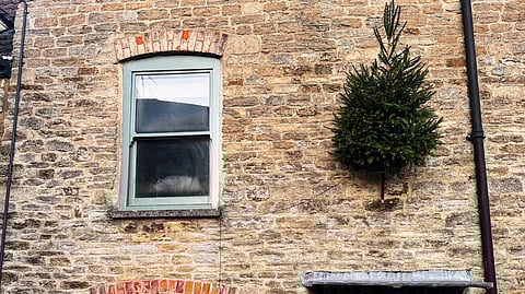Stone house with two green-framed windows. A small Christmas tree is mounted upside down on the wall. The green door has a wreath. Cozy and festive atmosphere.