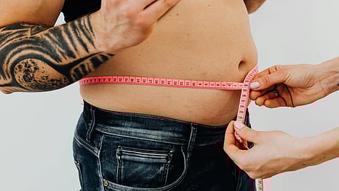 Man with tattoos holding a burger, while another person measures his waist with a pink tape measure; juxtaposes fast food and body awareness.