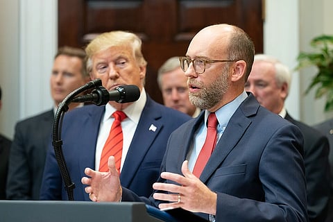 Vought in a suit and red tie speaks at a podium with a microphone, gesturing with his hands. Donald Trump stands behind, looking on. The setting is formal with other people blurred in the background.