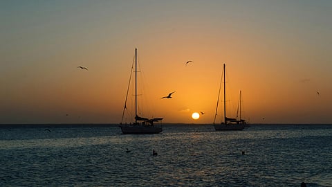 Sunset over the ocean with two silhouetted sailboats and birds flying. The sky is gradient orange to blue, creating a serene and peaceful scene.