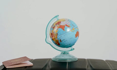 A small globe rests on a black suitcase, with a passport nearby. The scene evokes travel and exploration. The background is a simple, plain white.