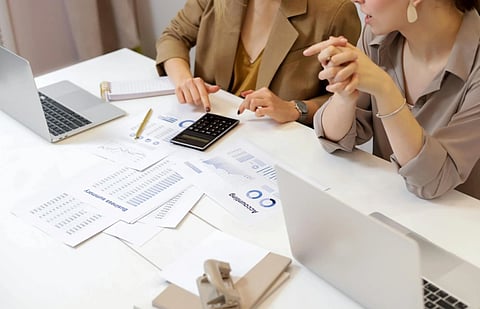 Two professionals are sitting at a table with laptops, papers, and a calculator. They appear to be discussing financial documents with focus and engagement.