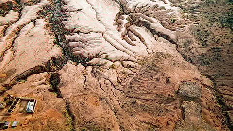 Aerial view of a barren, eroded landscape with deep ruts and ridges. Sparse vegetation and scattered buildings are visible, conveying isolation and ruggedness.