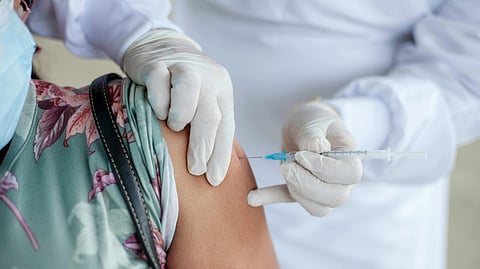 A healthcare worker in white protective clothing administers a vaccine to a person’s arm. The patient wears a floral shirt and face mask.