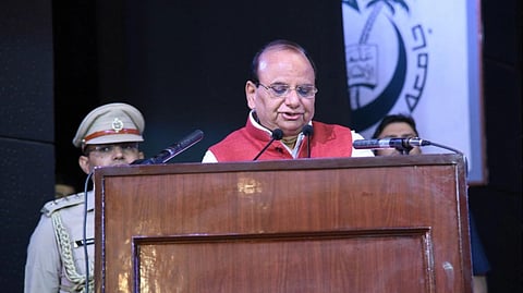Delhi LG V.K. Saxena in a red vest speaks at a wooden podium with microphones, flanked by a uniformed officer. The backdrop shows an abstract logo.