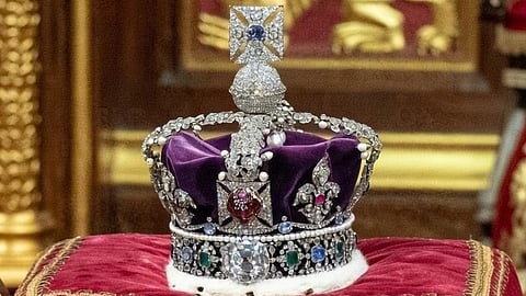 Ornate crown adorned with jewels sits on an intricately embroidered red velvet cushion, set against a backdrop of rich gold and red tapestry, conveying royalty.