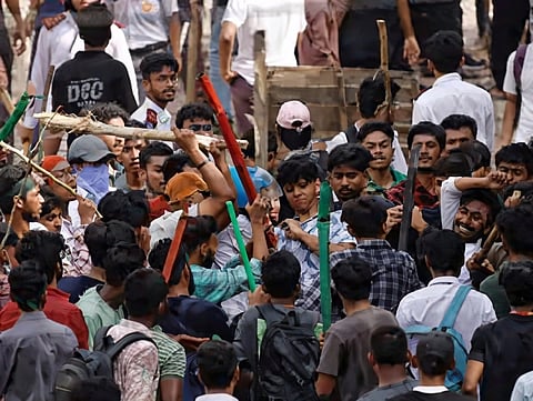 A large group of people, mostly young men, are engaged in a street altercation. Many hold colorful sticks, creating a tense, chaotic atmosphere.