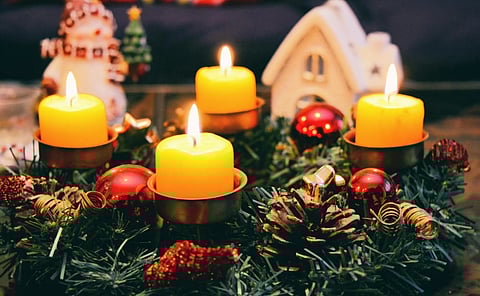 A festive Advent wreath with four glowing yellow candles, red baubles, and pinecones. In the background, a small house figurine and a snowman ornament add a cozy holiday feel.