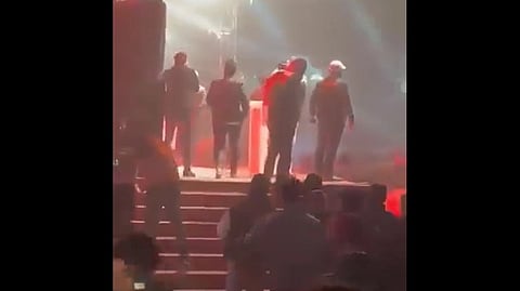 A group of people walking onto a brightly lit stage with red and white lighting, vandalizing the venue before the concert of the Bangladeshi rock musician James.
