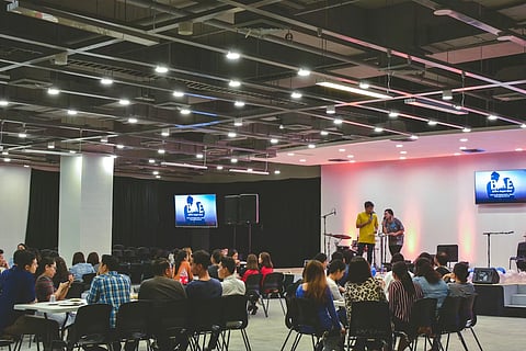 The image shows a hall where an event is taking place. It is a corporate event, people are sitting in groups, at seperate tables. One male and one female are hosting the event. There are lights above, screens on the walls.