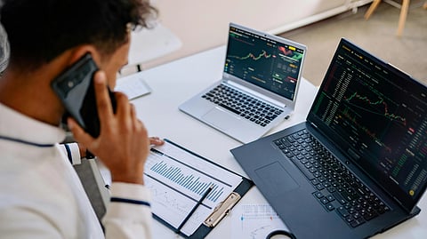 A person in an office talks on the phone while analyzing financial charts on two laptops and a clipboard, conveying a busy workplace atmosphere.