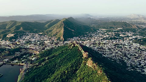 Aerial view of a lush green Aravalli Range with a historic fort wall stretching across the peaks in Jaipur, overlooking a sprawling city with densely packed buildings.