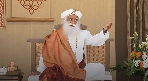 Sadhguru with a long white beard, wearing a white turban and robe with a brown shawl, gestures while seated in a calm room with flowers.