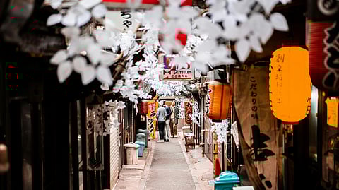 Narrow, bustling Japanese alleyway adorned with hanging white lanterns and decorations. People walk at the end, creating a cozy, lively atmosphere.
