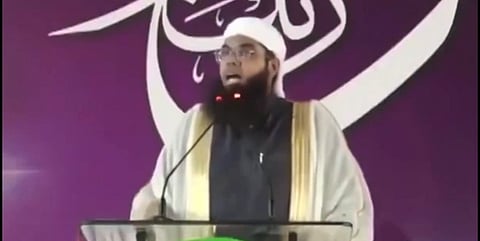 Mufti Nadwi in a white cap and robe speaks passionately at a podium with microphones. A purple backdrop with white Arabic calligraphy is visible behind him.