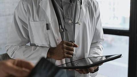 A doctor in a white coat and stethoscope holds a tablet, focused and attentive. Another blurred hand with a device is in the foreground, suggesting interaction.