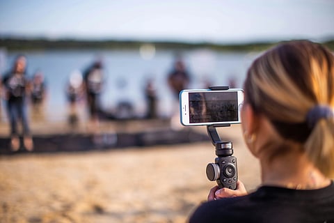 A woman films a sandy lakeside scene with a smartphone on a stabilizer. The background is blurred, showing people and a calm lake under clear skies.