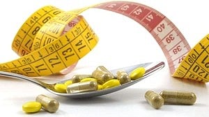 A spoon with yellow and gold capsules is in the foreground, surrounded by yellow and pink measuring tape, symbolizing diet pills and weight management.