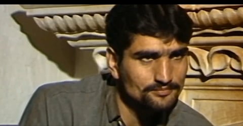 Bitta Karate with a serious expression is seated in front of an ornate, patterned wooden background. The atmosphere is contemplative and somber.