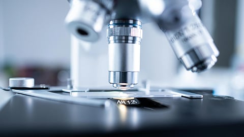 Close-up of a microscope with metallic lenses and an illuminated stage, set in a laboratory. The image conveys a sense of precision and scientific inquiry.