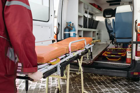 Person in a red uniform pushes an orange stretcher towards an open ambulance. Inside, medical equipment and seats are visible, creating an urgent tone.