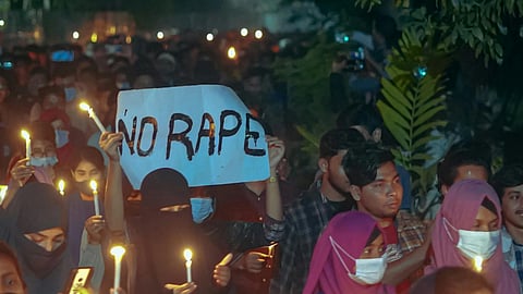 A crowd of people holding candles at a nighttime protest. A central sign reads "No Rape," conveying a message of solidarity and demand for justice.