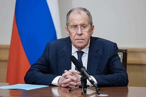The image shows Russian Foreign Minister Sergei Lavrov. He is addressing a press briefing, wearing a suit and tie, hands folded, sitting behind a mic. Behind him is the Russian Federation's flag.