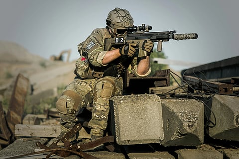 A soldier in camouflage and tactical gear, kneels on rubble, aiming a rifle. The setting is outdoors, suggesting a tense, active combat scene.